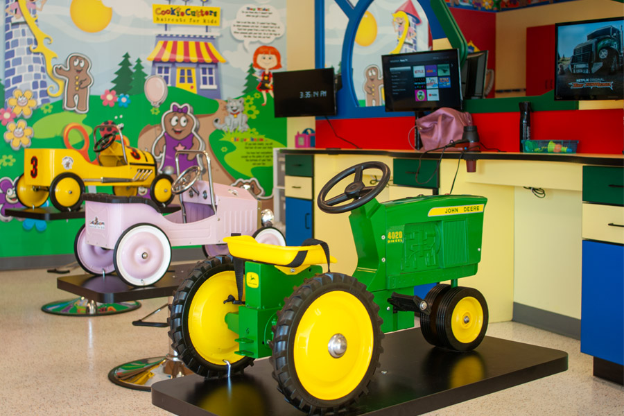 Photo of the interior of a Cookie Cutters salon with themed cutting chairs, TVs, and a mural on the wall.